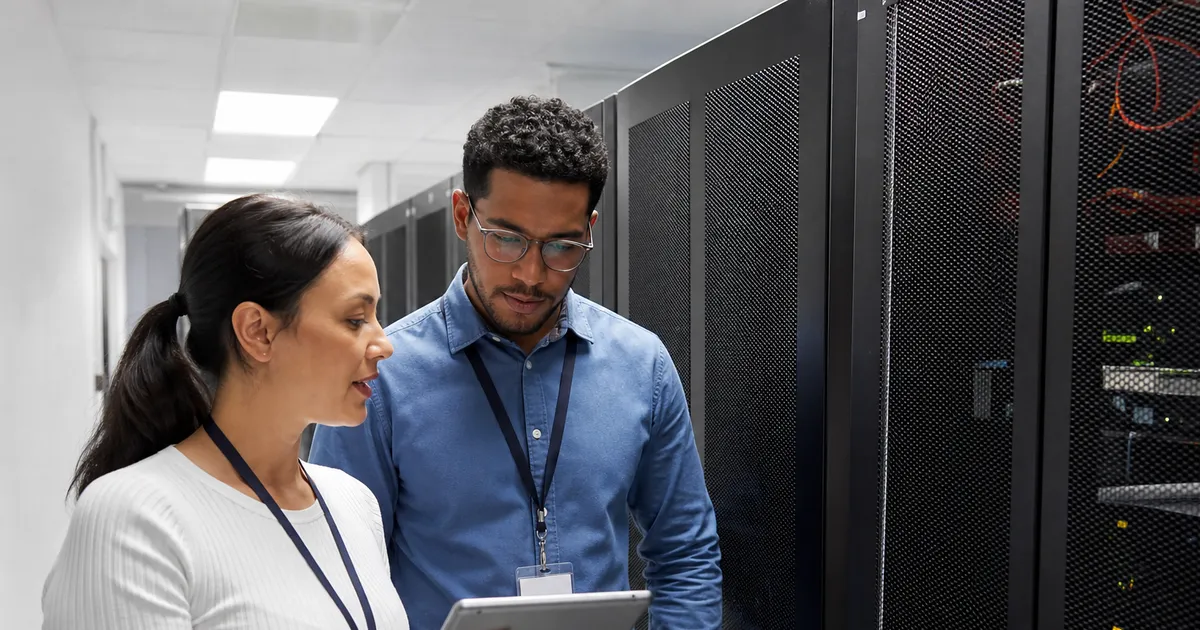 A technician works at a server rack in a dark data center, seen from behind with hands and shoulders in frame, cool blue light with warm amber accents